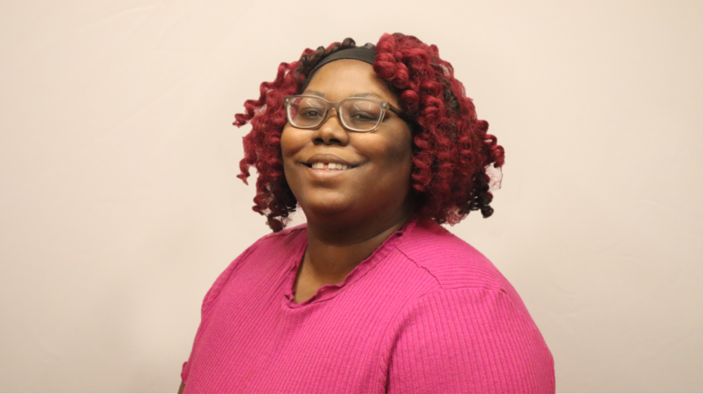 Professional headshot of a smiling woman with red curly hair and glasses, wearing a pink top against a light background